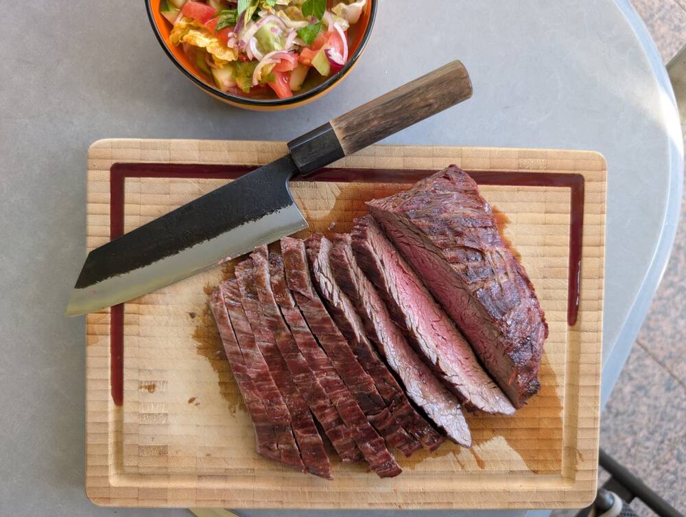 Sliced medium-rare flank steak on a wooden board, the knife still beside it and a bowl of tomato salad behind