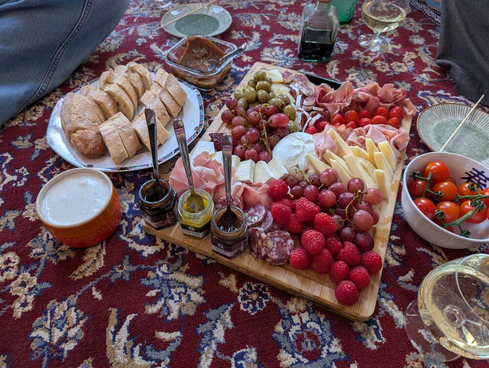 A picnic board on a red carpet with bread, cheese, cured ham, grapes and strawberries, a glass of beer at the edge