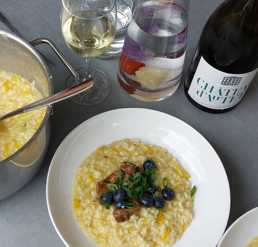 A plate of saffron risotto with mushrooms and blueberries, the pot and a glass of white wine beside it