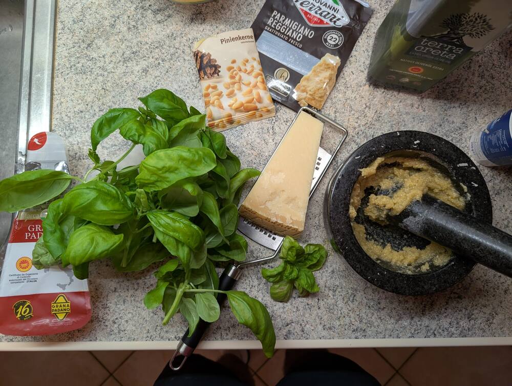 Pesto being ground in a stone mortar beside a bunch of basil and a wedge of parmesan