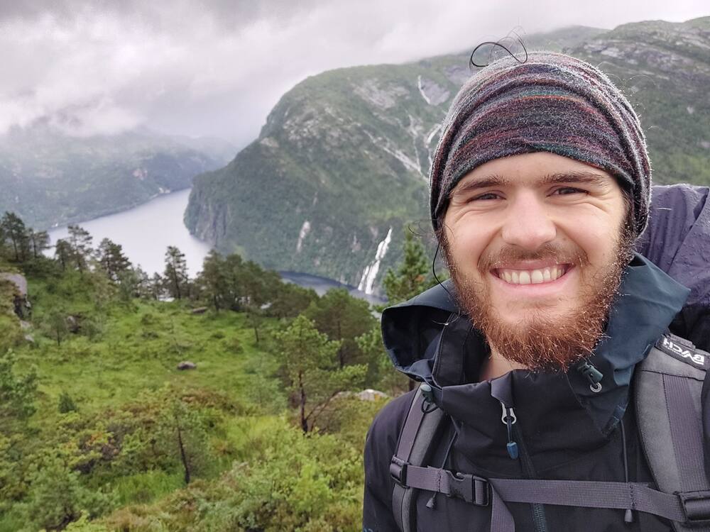 A selfie in rain gear on a forested slope, a fjord and its waterfalls below