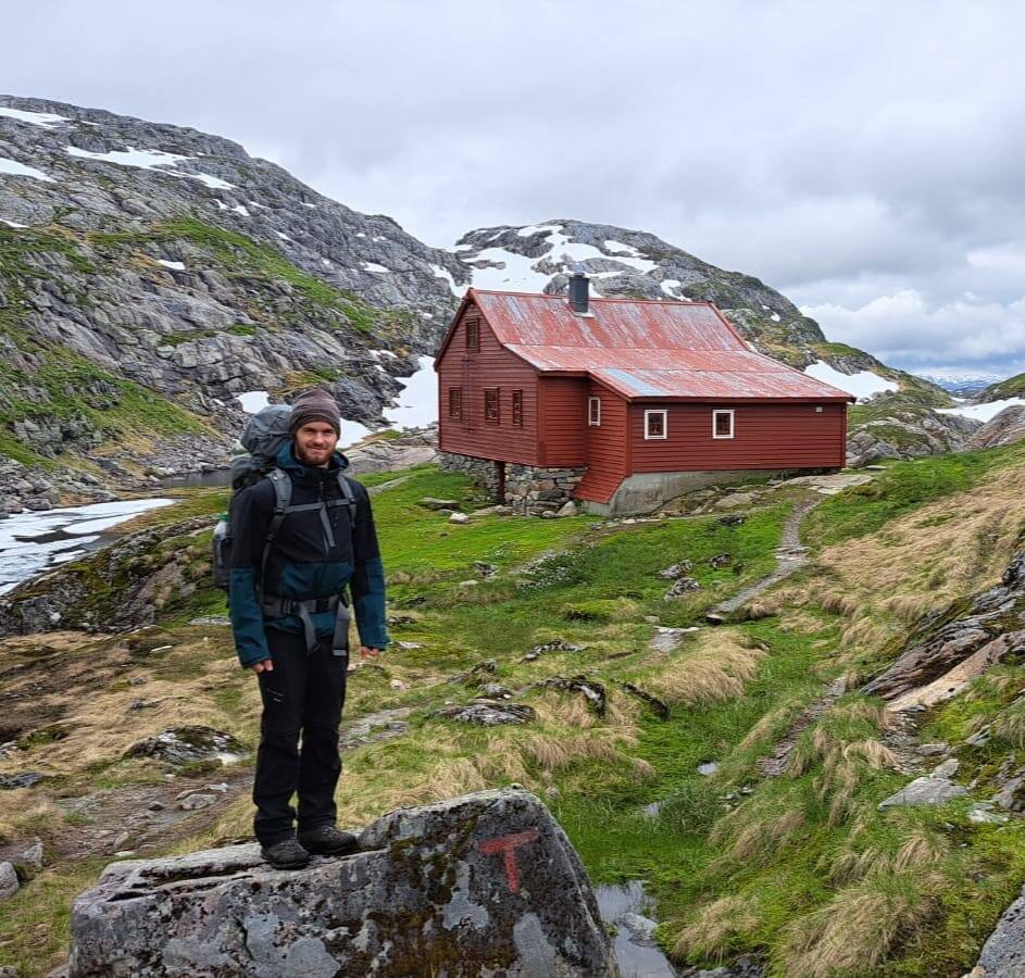 Standing with a backpack in front of a red mountain cabin among wet Norwegian rock