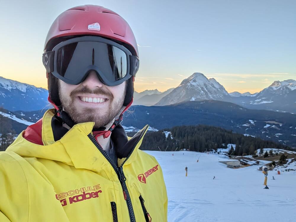 A ski selfie in helmet and goggles, the piste and distant peaks at dusk